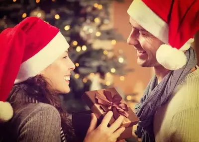 A couple in Santa hats exchanging Christmas gifts in front of a tree.